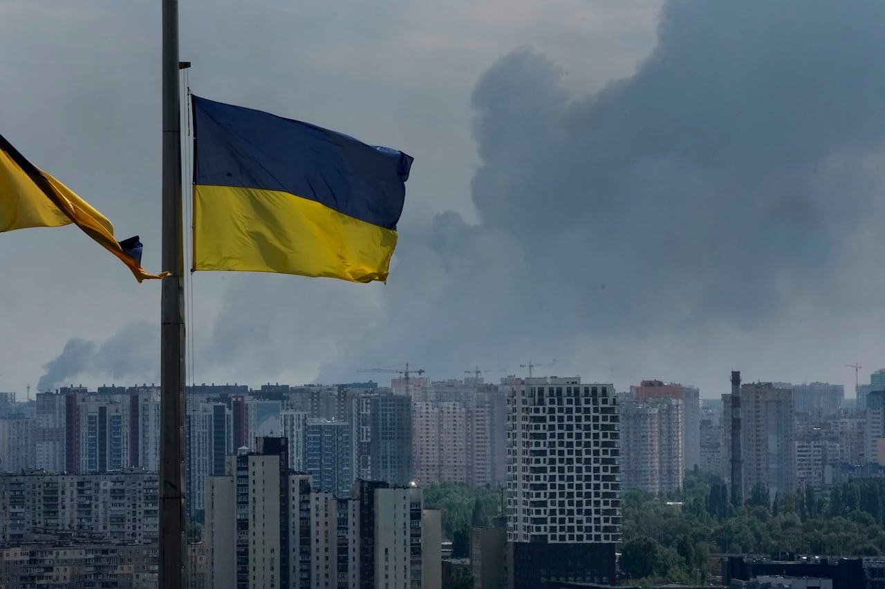 default-50 Ukrainian flags seen in foreground of image, with smoke rising above buildings in Kyiv, in the background.